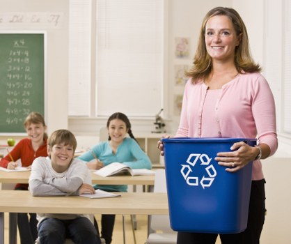 A woman standing in a classroom or training room holding a large blue plastic recycling bin with a white recycling symbol printed on the front. Behind her, children are seated at desks, smiling and looking towards the camera. The classroom features light-colored walls, a window with blinds, and a chalkboard with handwritten notes on the left side. The scene is brightly lit with natural and indoor lighting, creating a professional and organized environment that subtly emphasizes responsible waste management and recycling awareness relevant to local rubbish removal services in St Albans.