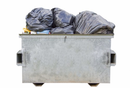 A large industrial skip bin made of galvanized metal, filled with multiple tightly packed black plastic garbage bags of various sizes, resting outdoors on a paved surface. The skip, featuring two side handles and a flat top edge, is positioned in front of a neutral background, possibly in a commercial or residential area within St Albans. The bags appear to contain general rubbish, with some showing slight creases and folds on their surface, while the metallic finish of the bin reflects ambient light, highlighting its utilitarian design. This scene exemplifies waste collection or rubbish removal activities that Commercial Waste St Albans might handle for local clients. The environment suggests an outdoor setting with minimal surrounding context, focusing on the bin's contents and structure, consistent with professional waste management services in the area.