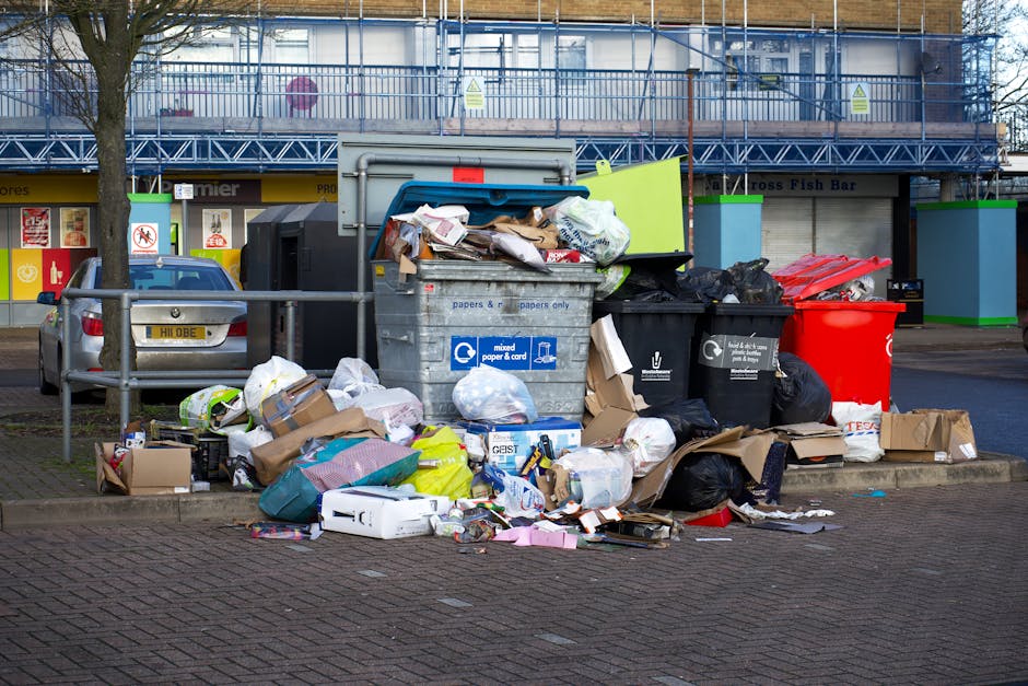 A street scene showing a waste collection vehicle with its rear open, revealing internal debris and waste materials. A worker wearing a high-visibility vest with red and yellow stripes is standing in 
