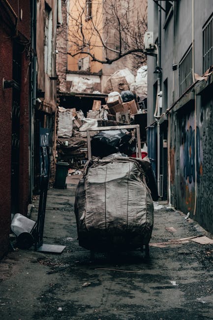 A person wearing a white glove and a wristwatch is seen lifting a large white woven polypropylene sack filled with garden debris, including green ivy and small twigs. The debris appears to consist of