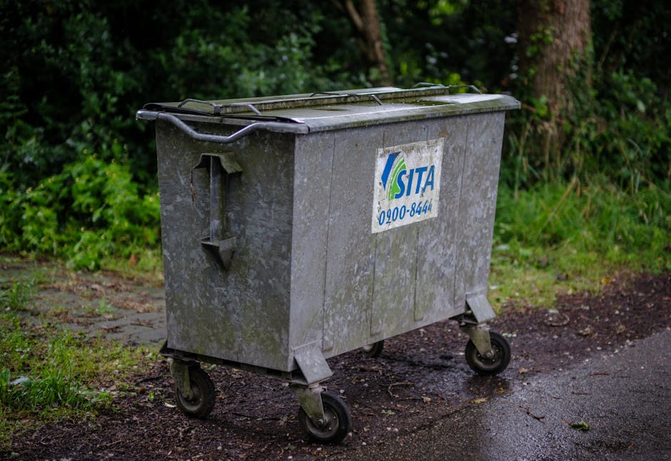 A large, wheeled metal rubbish bin positioned on a patch of ground next to a paved pathway, with a background of green foliage including bushes and trees. The bin is constructed from galvanized steel 