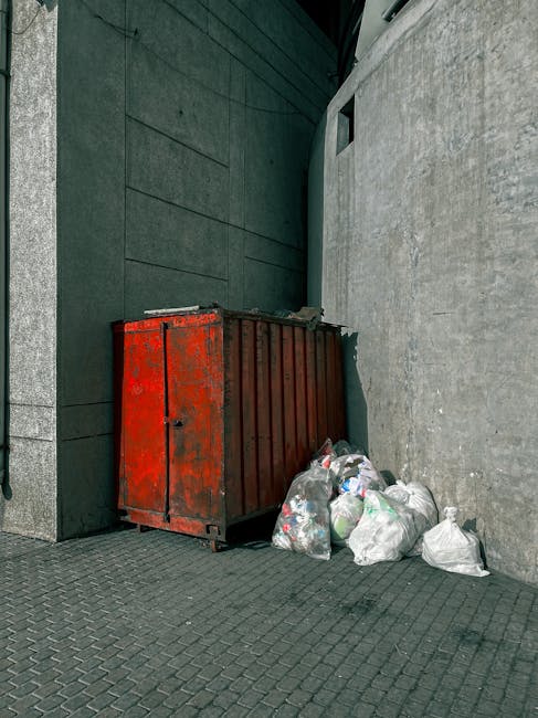 A large, rectangular metal rubbish bin with a slightly weathered, rough texture and a dull grey finish, positioned on a paved surface outdoors. The bin has four black caster wheels at the base for mob
